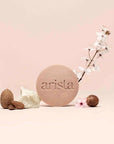Round pale pink shampoo bar for curly hair embossed with arista logo beside blossoms