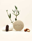 Beige round shampoo bar with arista branding, surrounded by olive branches and smooth stones