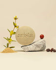 Round pale green arista shampoo bar with embossed logo beside yellow blossoms and a red apple on rocks