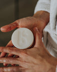 Hands hold a round white shaving bar with a towel in the background