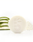 Round white shaving bar with a leaf imprint beside stacked aloe slices and a powder mound