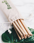 Natural bamboo drinking straws in a drawstring bag placed on a marble surface with green leaves