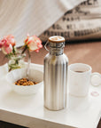 Close-up of a stainless steel water bottle with a wooden lid and metal handle on a white table with a mug and cereal bowl nearby