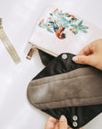 Embroidered beige zip bag on a white surface with a hand holding a black pouch nearby