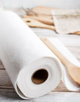 Close up of a white reusable paper towel roll on a rustic wooden surface with a sheet partially unrolled and a wooden spoon nearby.