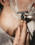 Person shaving the jawline with a bamboo safety razor and foamy lather on the face.