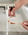 Copper tongue scraper being rinsed under running water from a metal faucet