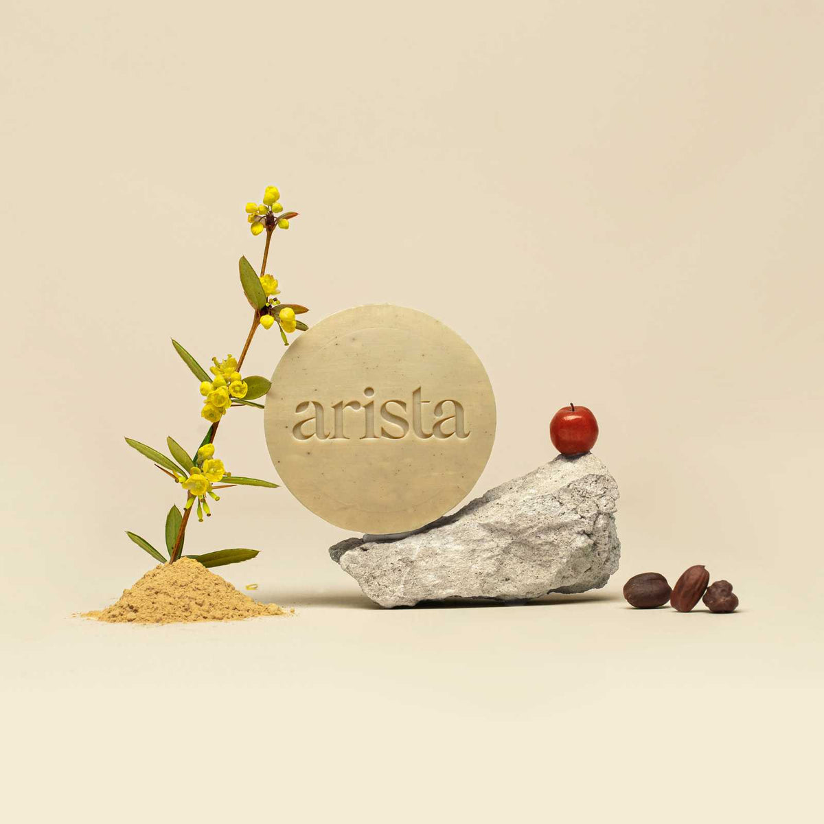 Round pale green arista shampoo bar with embossed logo beside yellow blossoms and a red apple on rocks