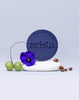 Purple arista circular shampoo bar with embossed logo on a white surface surrounded by a purple flower and green grapes