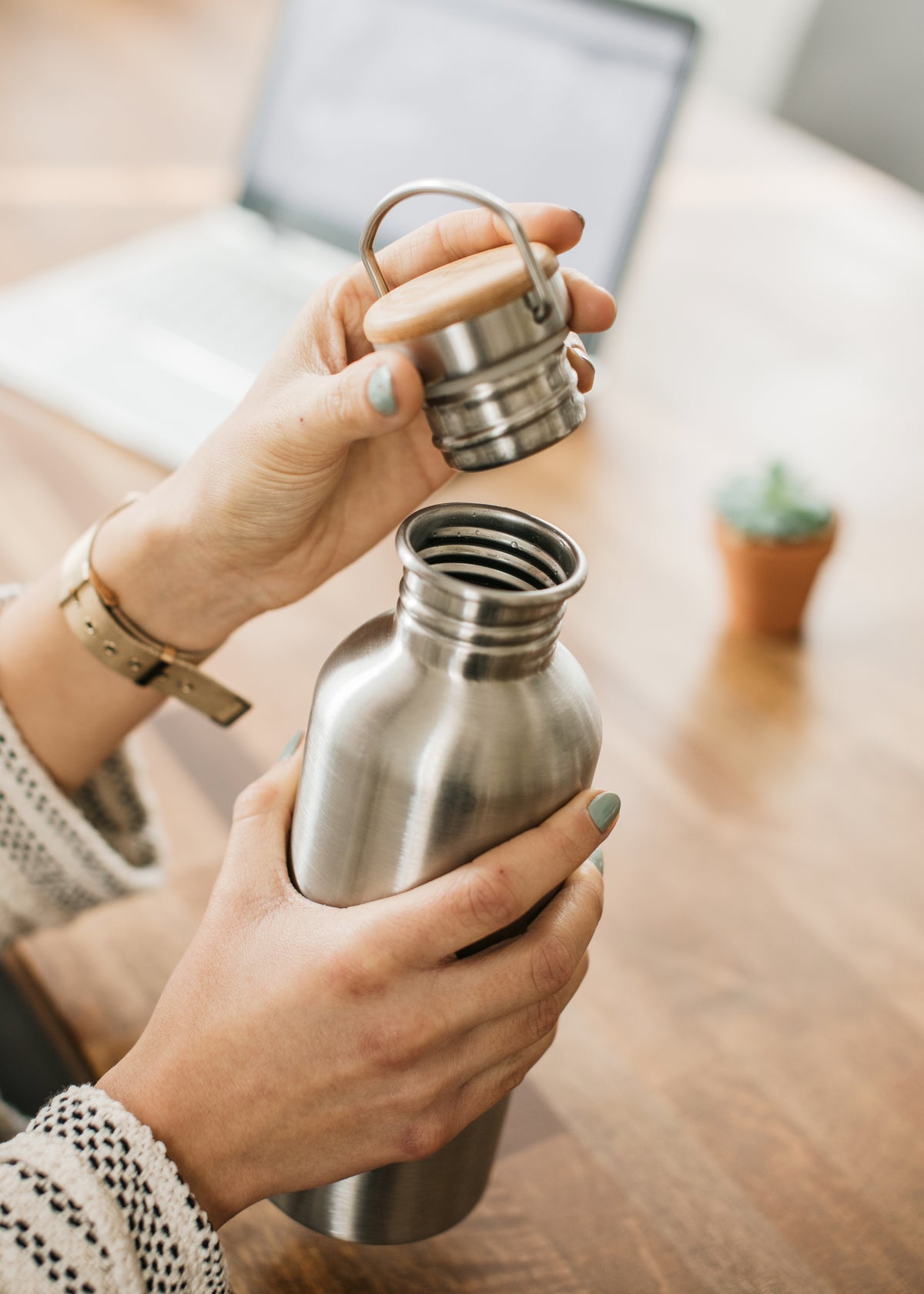 Person holding a bambaw stainless steel water bottle in natural steel colour with a wooden cap indoors.