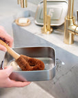 Hand holding a wooden dish brush with brown bristles over a metal container on a kitchen counter.