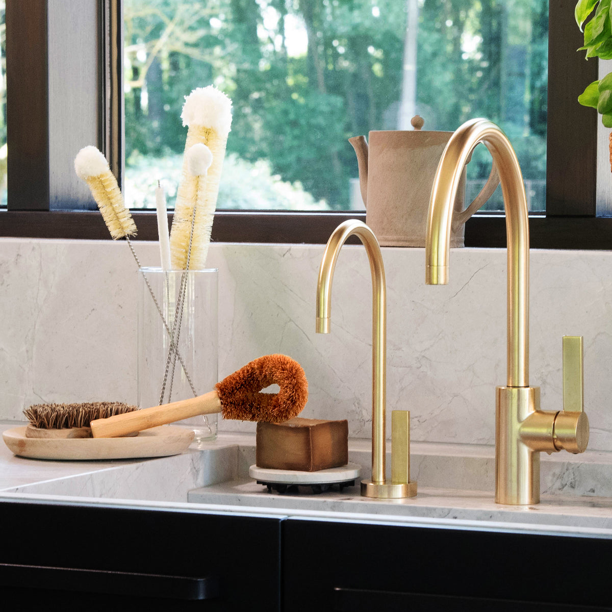 Cream fluffy dish brushes in a glass holder on a marble counter by a window in a bright kitchen.