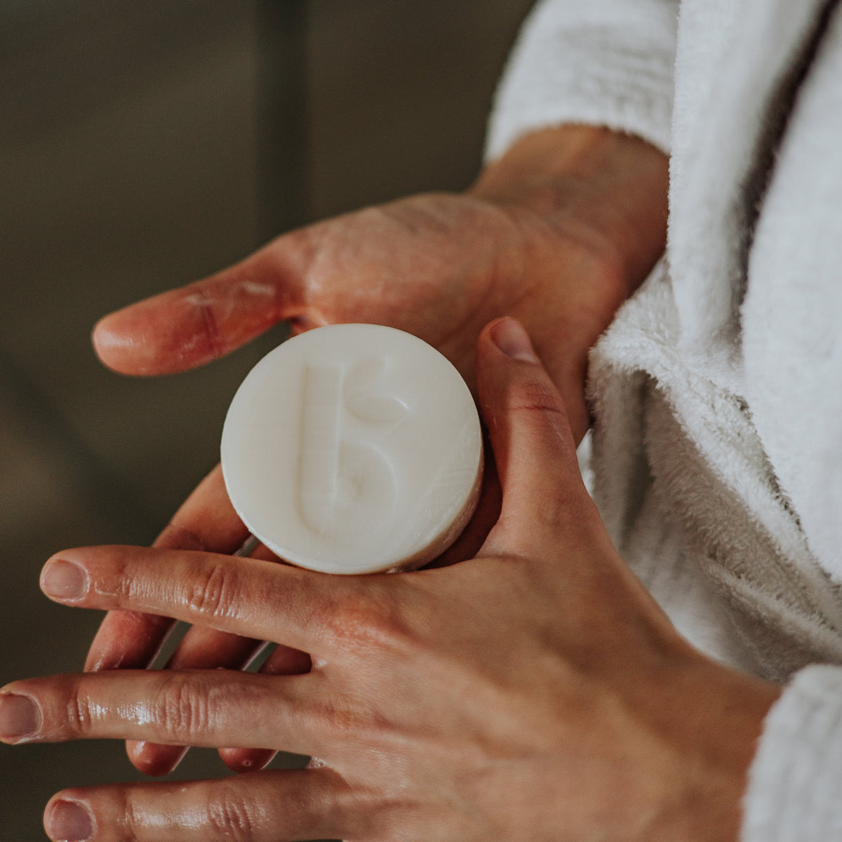 Hands hold a round white shaving bar with a towel in the background