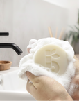 Hand holding a bambaw shaving soap bar of soap with lather, in front of a sink with a black faucet.