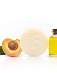 Round ivory shaving soap with a peach half and a bottle of oil on a white background