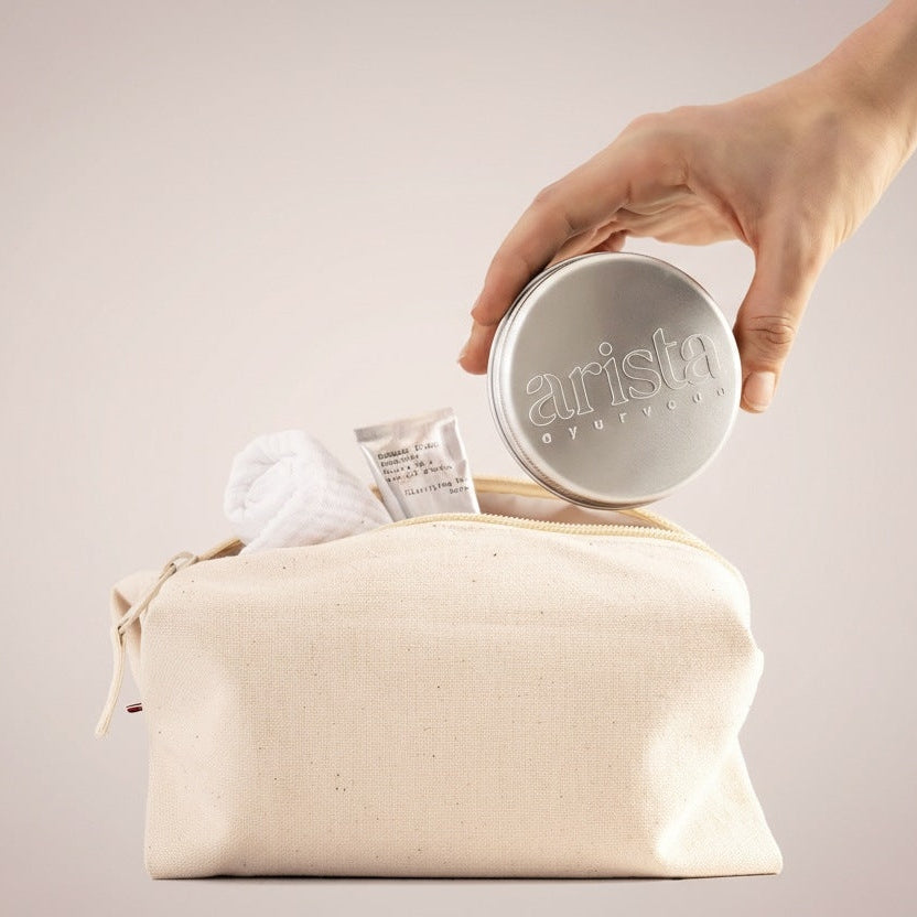 Hand placing a round aluminium tin labeled Arista into a beige canvas toiletry bag with a towel and sample packet nearby.