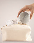 Hand placing a round aluminium tin labeled Arista into a beige canvas toiletry bag with a towel and sample packet nearby.
