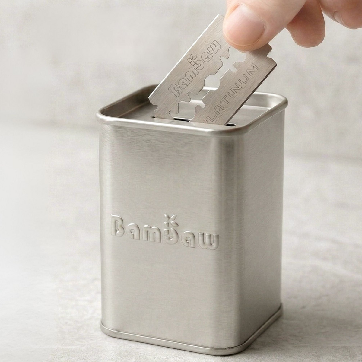 Metallic razor blade being inserted into a metal Bambaw blade bank container on a light gray background