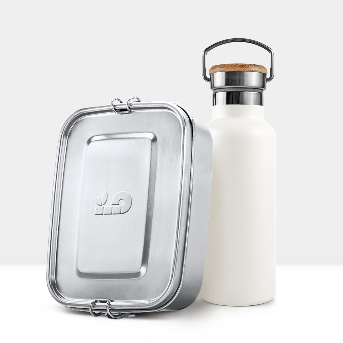 Silver metal lunch tin with hinged clasp beside a white insulated bottle topped with a cork lid and metal handle.