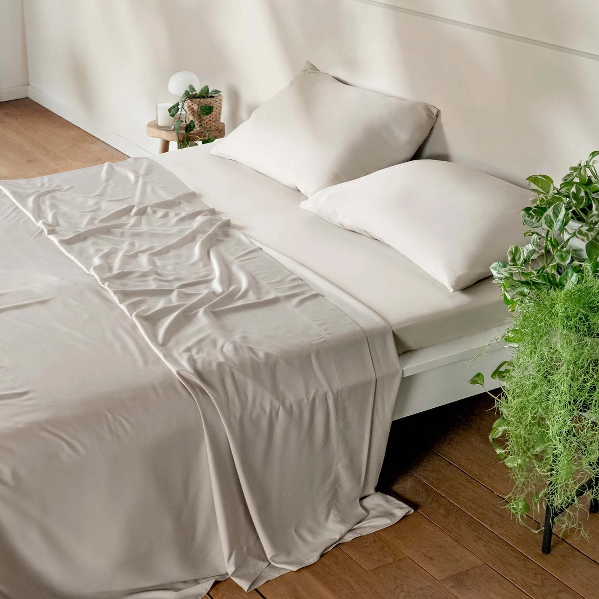 A neatly made bed with light beige bambaw cozy bamboo bedding, including a flat sheet, fitted sheet and two pillows, styled in a minimalist bedroom with a wooden floor, a side table with a plant, and a hanging green plant in the foreground.