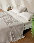 A neatly made bed with light beige bambaw cozy bamboo bedding, including a flat sheet, fitted sheet and two pillows, styled in a minimalist bedroom with a wooden floor, a side table with a plant, and a hanging green plant in the foreground.