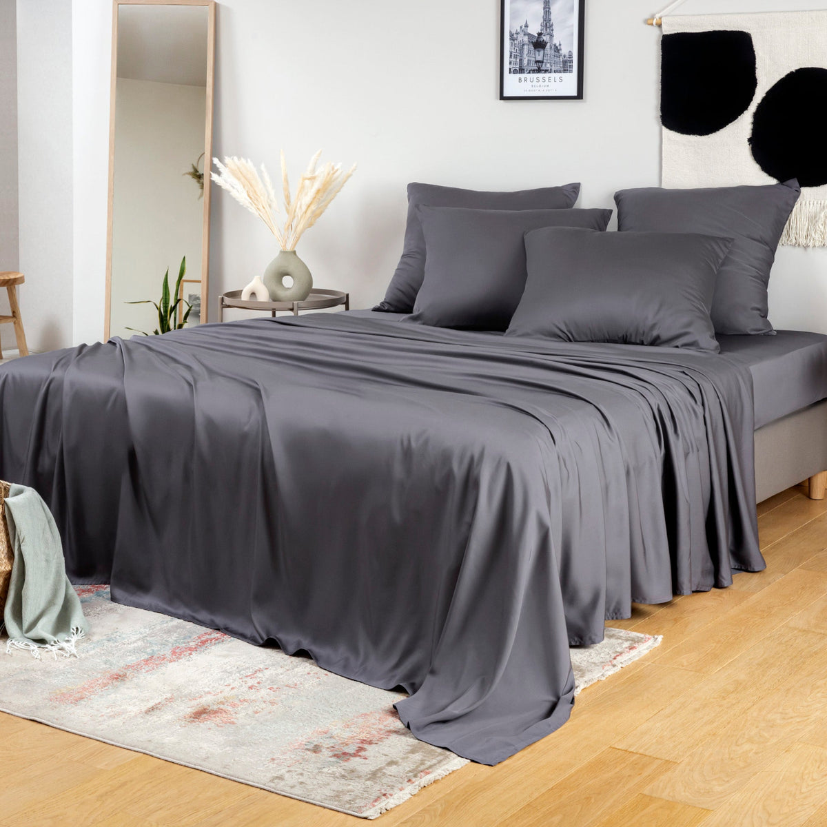 A bed dressed in charcoal bambaw cozy bamboo bedding bedding, including a sheet, duvet, and matching pillowcases, styled in a bright bedroom with a mirror, side table, framed wall art, and light wood floor.