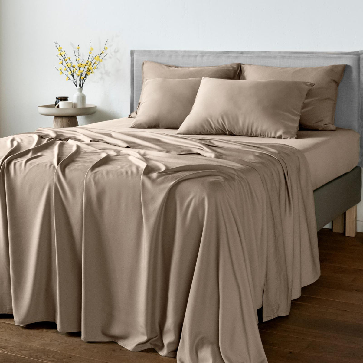 A neatly made bed with a smooth taupe-beige bamboo bedsheet set, including a fitted sheet and matching pillowcases, styled in a modern bedroom setting with a gray upholstered headboard and a small side table with yellow flowers.
