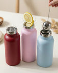 Three pastel insulated bottles on a white surface, lemon in the middle and a tea infuser hovering above the right bottle.