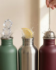 Three stainless steel water bottles in green silver and burgundy with wide mouths ice and a lemon slice at the openings and a tea infuser being added