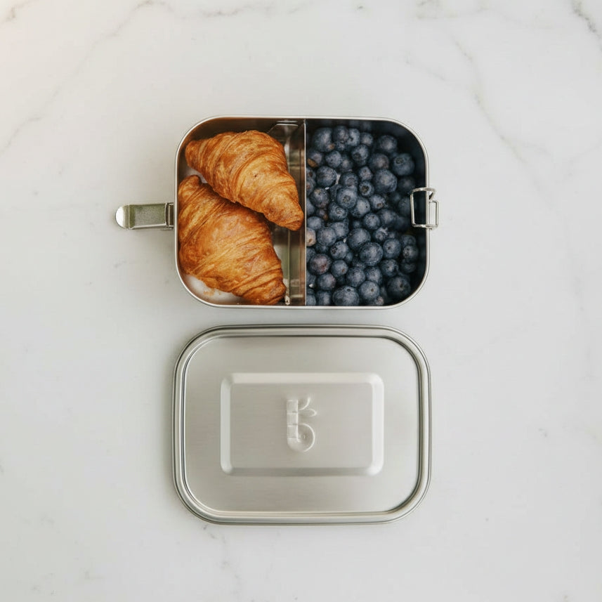 A bambaw stainless steel lunch box shown open on a white marble surface, with two croissants in one compartment and blueberries in the other. The matching lid is placed below the box.
