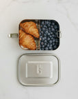 A bambaw stainless steel lunch box shown open on a white marble surface, with two croissants in one compartment and blueberries in the other. The matching lid is placed below the box.