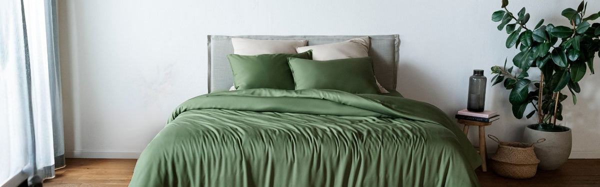 Bed with olive green bedding in a room with a plant and side table.