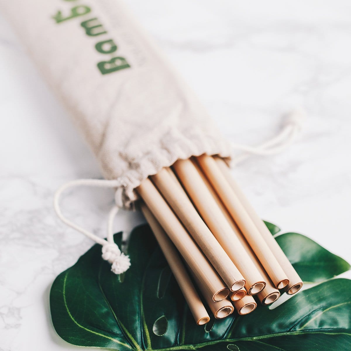 Natural bamboo drinking straws in a drawstring bag placed on a marble surface with green leaves
