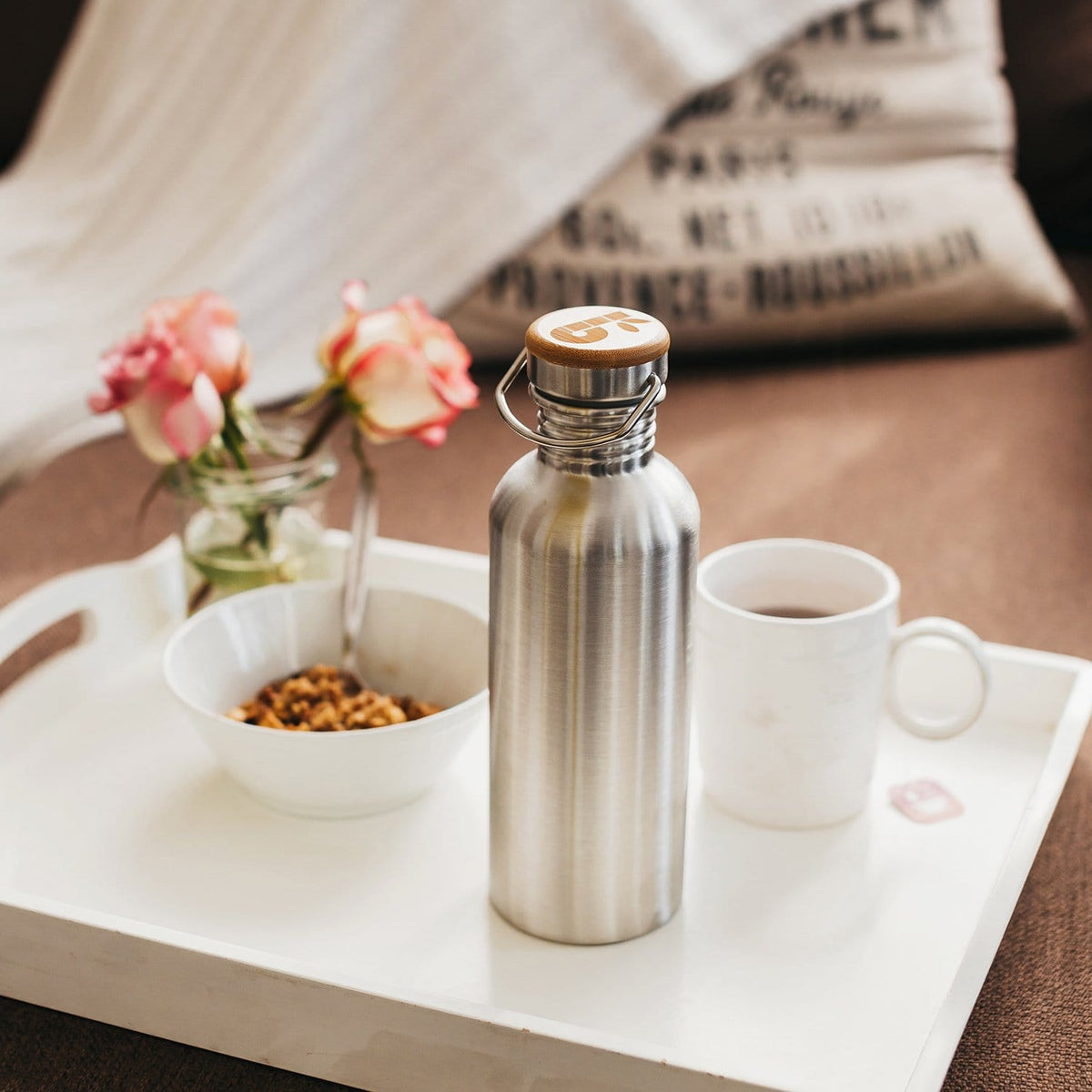 Close-up of a stainless steel water bottle with a wooden lid and metal handle on a white table with a mug and cereal bowl nearby