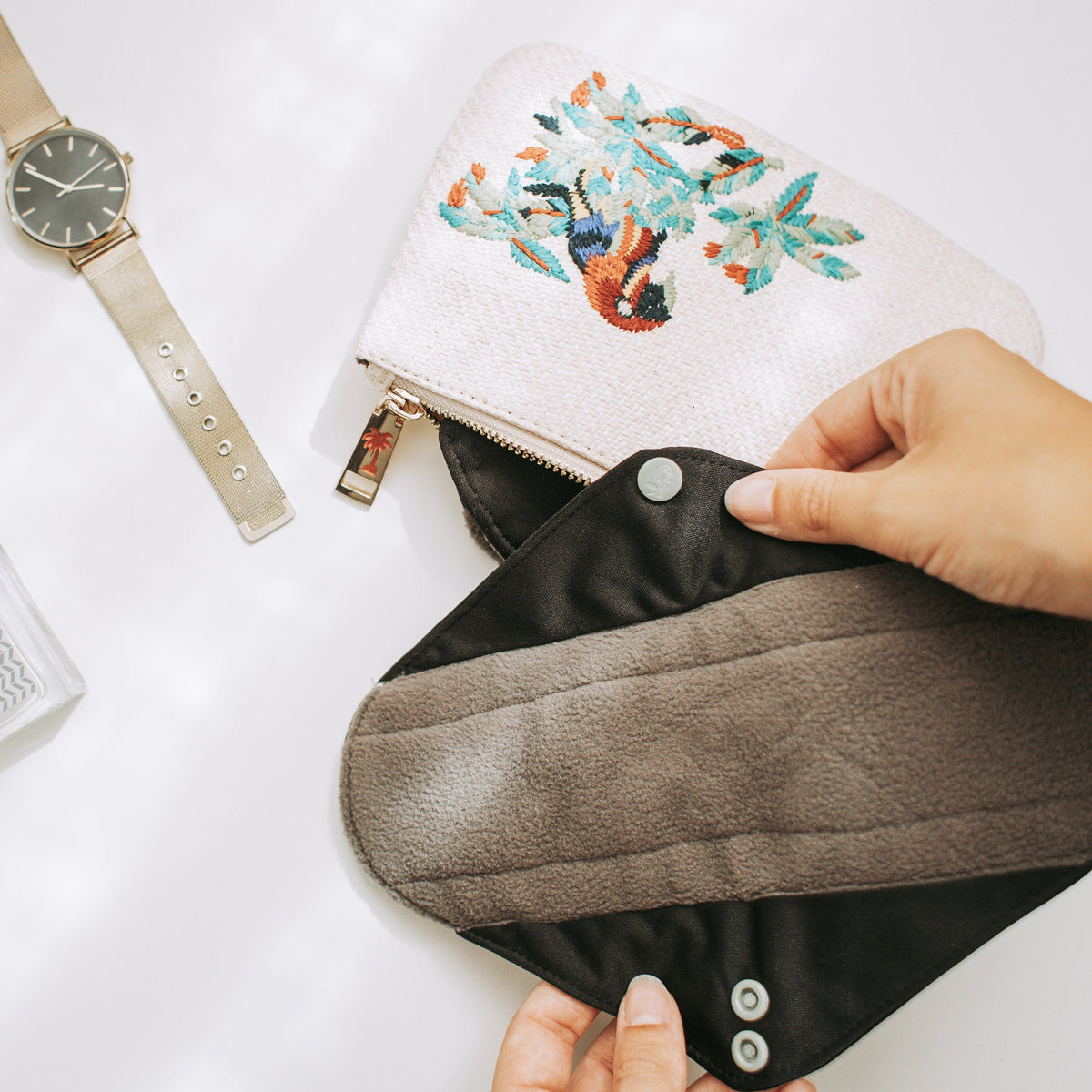 Embroidered beige zip bag on a white surface with a hand holding a black pouch nearby