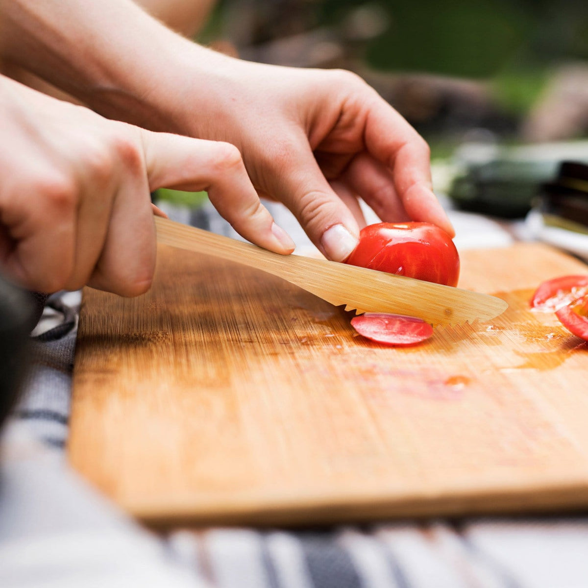Hands slice a tomato with a bamboo knife on a wooden cutting board
