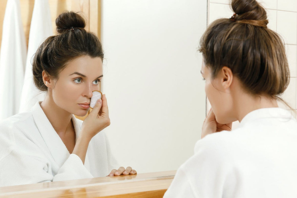 Woman in a white robe applies a reusable makeup remover pad to her face in a bathroom mirror.
