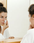 Woman in a white robe applies a reusable makeup remover pad to her face in a bathroom mirror.