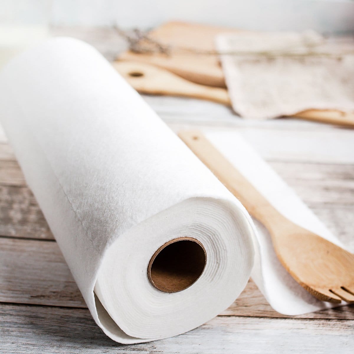 Close up of a white reusable paper towel roll on a rustic wooden surface with a sheet partially unrolled and a wooden spoon nearby.