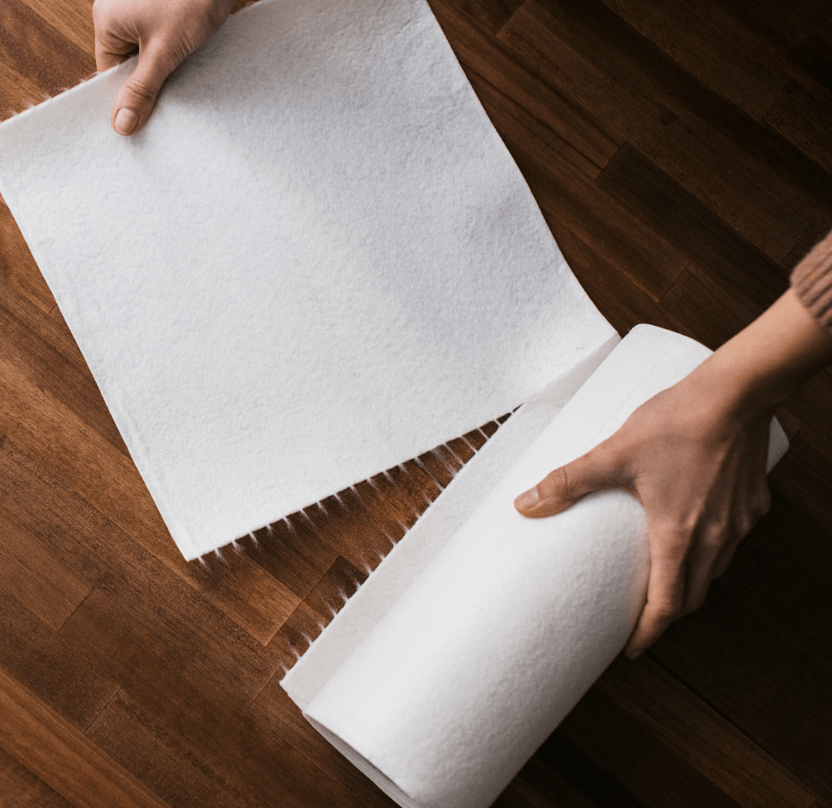 Hands unrolling a sheet from a reusable paper towel roll onto a wooden floor.