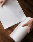 Hands unrolling a sheet from a reusable paper towel roll onto a wooden floor.