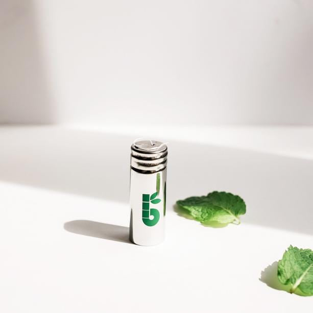 Close-up of a small metal cap with a green logo beside mint leaves on a white surface