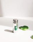 Close-up of a small metal cap with a green logo beside mint leaves on a white surface